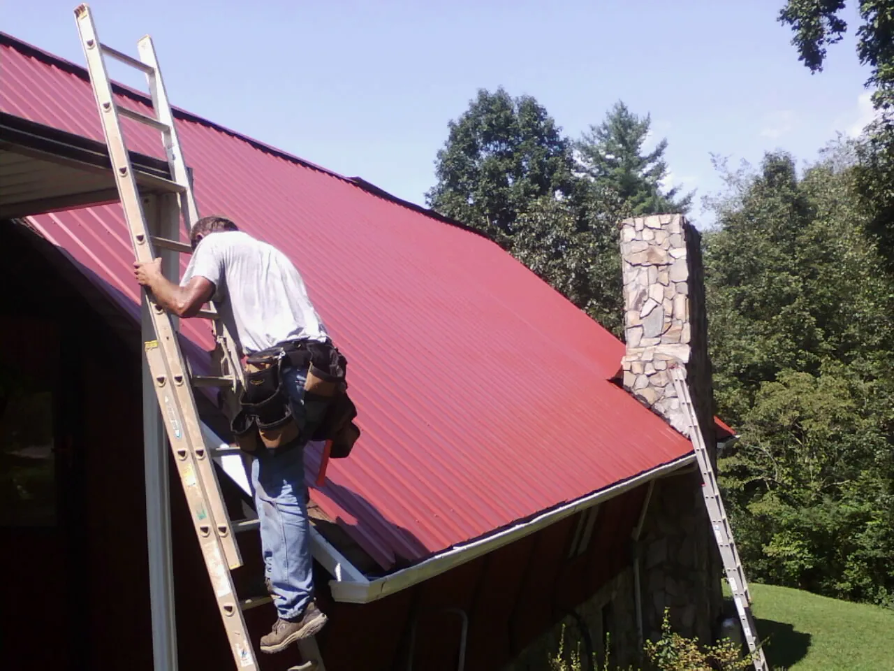 Roofer on a ladder beside a red metal roof and stone chimney — Soffit & Fascia Repair by Roofing Brothers in Clark Fork